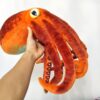 Une peluche de poulpe orange avec des ventouses brunes. Le poulpe est tenu par une main.