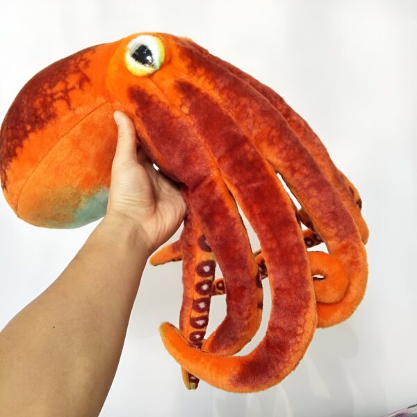Une peluche de poulpe orange avec des ventouses brunes. Le poulpe est tenu par une main.