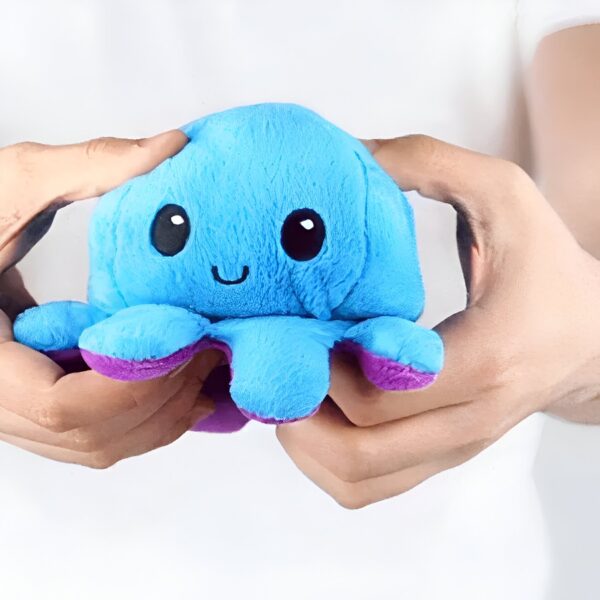 peluche-pieuvre-reversible-emoji-heureux-en-colere-cadeau-enfant-5a Une peluche de pieuvre bleue et violette avec un visage souriant, tenue dans les mains.