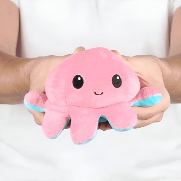 peluche-pieuvre-reversible-emoji-heureux-en-colere-cadeau-enfant-7a Une main tenant une peluche rose en forme de pieuvre avec des yeux noirs et un sourire. Les tentacules de la pieuvre sont bleu clair.