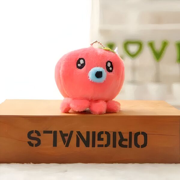 Une peluche rose en forme de pieuvre avec de grands yeux noirs et un nez bleu, assise sur un coffret en bois avec l'inscription "ORIGINALS".