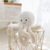 Une peluche blanche en forme de pieuvre souriante avec des tentacules beige sur une table en métal blanc.