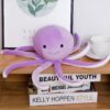 Une peluche de poulpe violette avec un sourire sur un étagère en bois avec des livres.