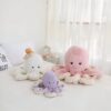 Trois peluches poulpes : une blanche avec un petit ours en peluche sur la tête, une rose et une violette. Les peluches sont disposées sur un lit blanc.