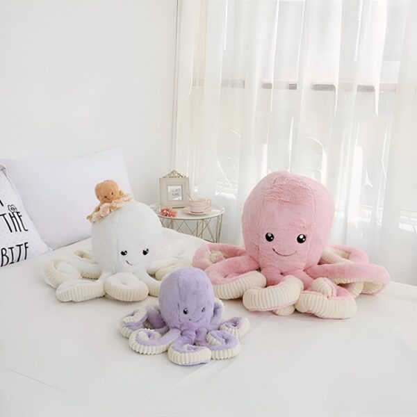 Trois peluches poulpes : une blanche avec un petit ours en peluche sur la tête, une rose et une violette. Les peluches sont disposées sur un lit blanc.