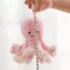 Une main tenant un poulpe rose en peluche, mesurée avec un ruban à mesurer. La peluche est environ 16 cm de haut.