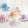 Cinq peluches poulpes de différentes couleurs sont disposées sur un lit blanc. Les poulpes sont souriants et ont des yeux noirs.