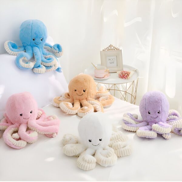 Cinq peluches poulpes de différentes couleurs sont disposées sur un lit blanc. Les poulpes sont souriants et ont des yeux noirs.