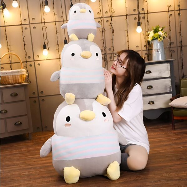 Une jeune femme souriante assise sur un sol en bois, tenant une pile de trois peluches pingouins grises et blanches rayées.