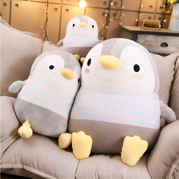 Trois peluches de pingouins, portant des rayures roses et bleues, se blottissent sur un canapé en tissu beige.
