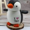 Un adorable peluche de pingouin blanc et noir avec un bec orange et des pieds orange, debout sur un socle en bois. Il a une inscription "Je t'aime" sur la poitrine.