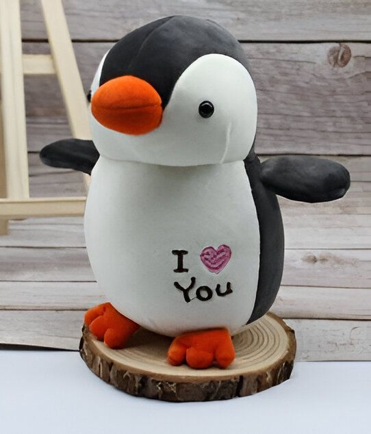 Un adorable peluche de pingouin blanc et noir avec un bec orange et des pieds orange, debout sur un socle en bois. Il a une inscription "Je t'aime" sur la poitrine.