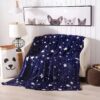 peluche-plaid-etoiles-lumineuses-bleu-nuit-5a-1 Une couverture bleu foncé avec des étoiles blanches et des traînées d'étoiles filantes sur un canapé blanc dans une pièce décorée avec des lumières et des décorations.