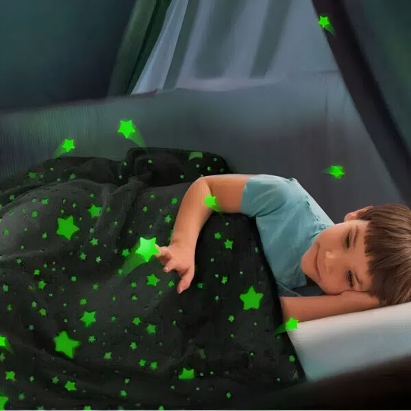 peluche-plaid-etoiles-lumineuses-cadeau-parfait-pour-enfants-3a-2 Un enfant dort sous une couverture noire parsemée d'étoiles vertes qui brillent dans le noir.