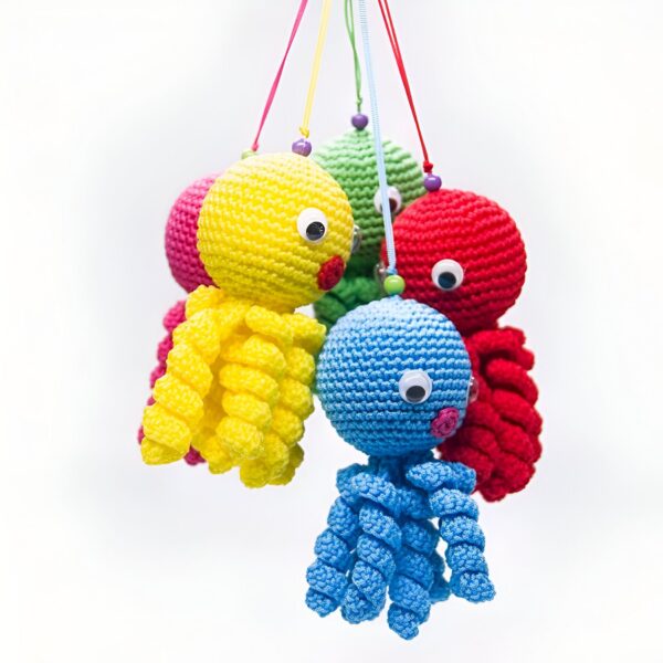 Jouet de bébé en forme de pieuvre crocheté dans des couleurs vives, suspendu à un mobile. Le jouet est fait de fils jaunes, rouges et bleus avec des yeux blancs et de petites perles colorées.