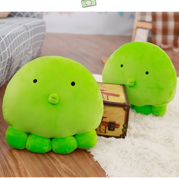 Deux peluches vertes en forme de pieuvre avec de grands yeux et un bec. Les peluches sont sur un sol en bois clair. L'une des peluches est devant un coffre en bois et l'autre est sur un tapis blanc.