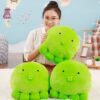 Une jeune femme tient une peluche verte en forme de pieuvre, deux autres peluches similaires sont posées à côté d'elle.