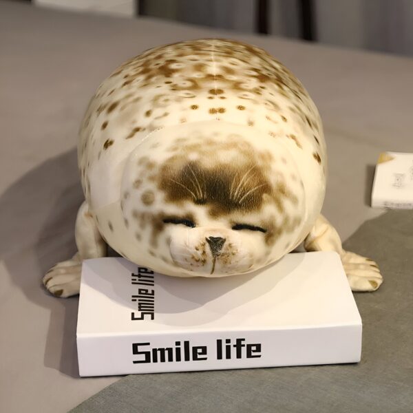 peluche-realiste-phoque-xxl-4a-1 Un gros coussin en forme de phoque avec des points marrons et une expression faciale mignonne. Le coussin est posé sur un livre blanc avec le texte "Smile life" écrit dessus.