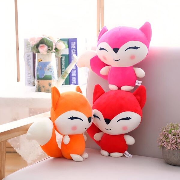 Trois peluches renards mignons, un rose, un rouge et un orange, assis sur un canapé.