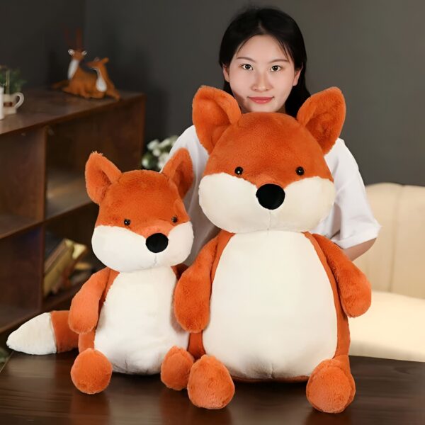 Deux peluches renard, une grande et une petite, sont posées sur une table. Une jeune femme sourit en tenant la grande peluche.