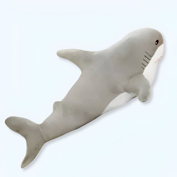 Une peluche en forme de requin gris avec une nageoire dorsale et une queue. Le requin a des yeux noirs et une bouche ouverte.