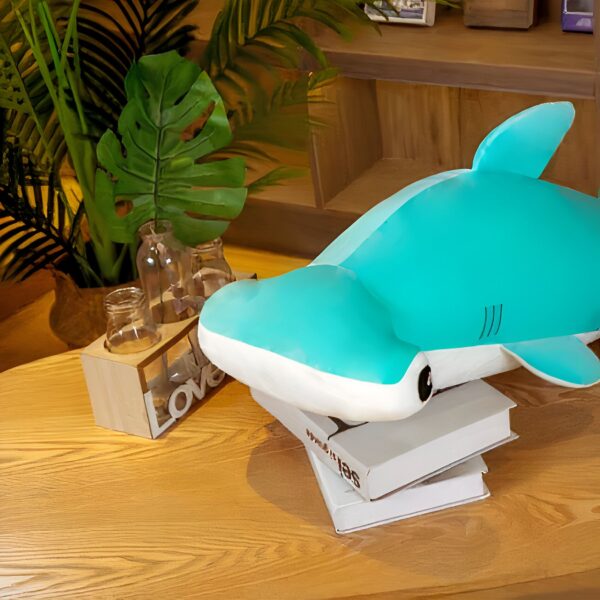 Une peluche de requin marteau bleu repose sur un livre sur une table en bois. Un vase en verre contient une plante verte.