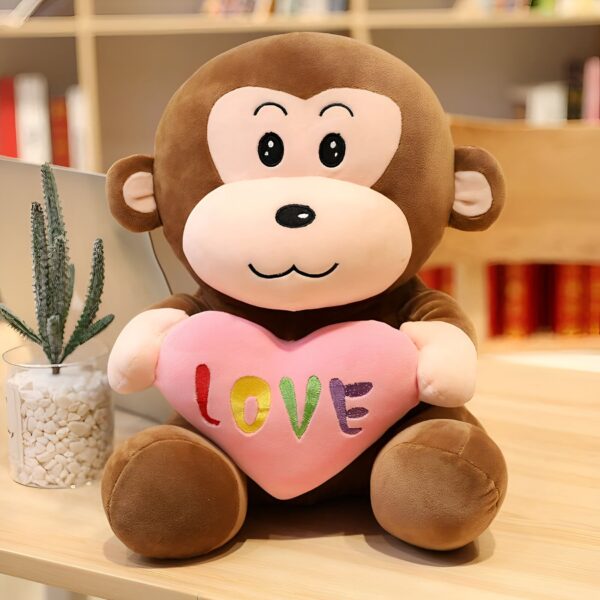 Une peluche de singe marron souriant tenant un cœur rose avec le mot "LOVE" écrit en arc-en-ciel.