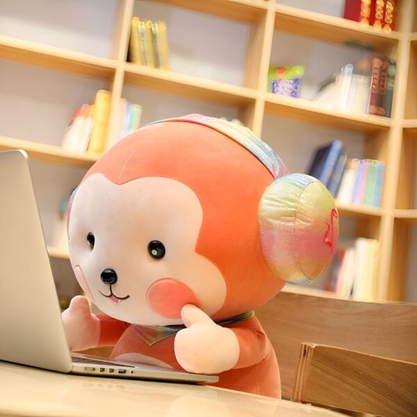 Une peluche de singe orange avec un casque arc-en-ciel est assis sur un bureau en bois en train d'utiliser un ordinateur portable.