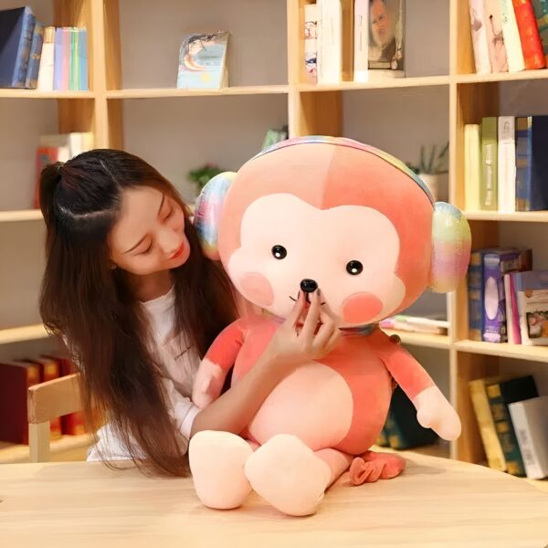 Une jeune femme souriante tenant une grande peluche de singe rose avec des oreilles arc-en-ciel.