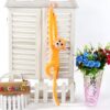 peluche-singe-kawaii-jaune-a-longue-queue-jouet-doux-pour-enfant-3a-2 Un singe en peluche jaune suspendu à une porte en bois blanc avec un vase de fleurs bleues et roses à côté.
