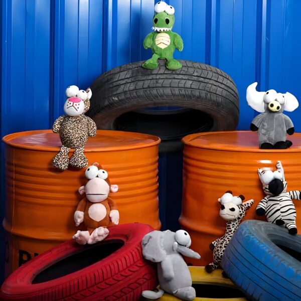 Des peluches amusantes avec de grands yeux, dont un crocodile vert, un singe brun, un zèbre noir et blanc, un éléphant gris, un girafe orange et un buffle gris, posées sur des barils et des pneus orange et bleus.