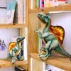 Deux dinosaures en peluche, un grand et un petit, sont assis sur une étagère en bois. Le grand dinosaure est vert avec une crête orange et jaune. Le petit dinosaure est vert avec une crête orange.