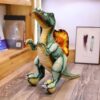 Peluche de dinosaure vert et jaune avec une crête orange sur le dos, debout sur un sol en bois.