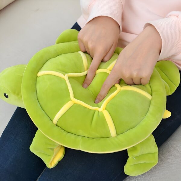 Une peluche verte en forme de tortue avec une carapace jaune, tenue par des mains.