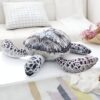 Une grande peluche en forme de tortue marine posée sur une table. La tortue est grise et blanche, avec une carapace détaillée.