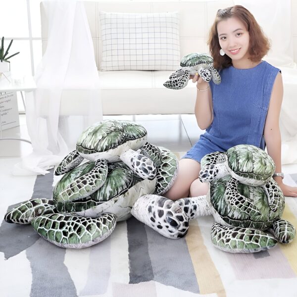 peluche-tortue-de-mer-verte-jouet-calin-pour-enfants-oreiller-doux-2a Une jeune femme souriante tient un coussin en forme de tortue. À ses pieds, il y a deux grands coussins en forme de tortue, un en haut de l'autre.