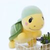 Une adorable tortue en peluche jaune avec une carapace verte, des yeux noirs et un sourire, posée sur un panier blanc.