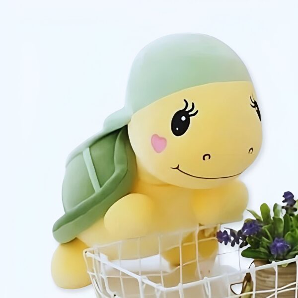 Une adorable tortue en peluche jaune avec une carapace verte, des yeux noirs et un sourire, posée sur un panier blanc.