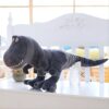 Un dinosaure en peluche gris se trouve sur un banc blanc. Il est souriant et a de grands yeux.