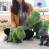Une femme assise sur un tapis blanc avec trois peluches de dinosaures verts et gris. Les dinosaures ont de grands yeux et des sourires amicaux.