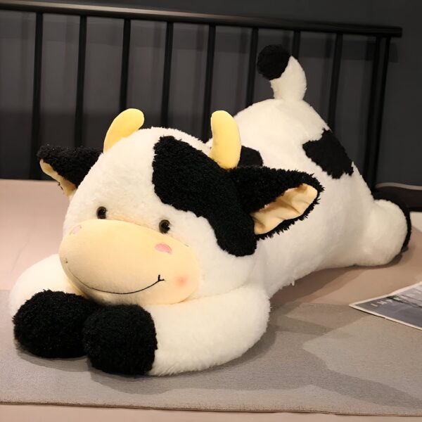 Une grosse peluche de vache blanche et noire avec des yeux marrons et un sourire. Elle est allongée sur un lit.