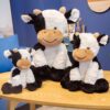 Trois adorables peluches en forme de vache, avec des taches noires et blanches, sont assises sur une table en bois. La plus grande vache est assise sur ses pattes arrière, tandis que les deux plus petites sont assises sur leurs pattes avant. Les trois peluches ont un air joyeux et souriant.