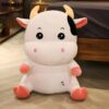 Une peluche blanche en forme de vache avec des détails roses et noirs. La vache a de grands yeux noirs, un sourire et une petite tache noire sur l'oreille. Elle est assise sur un plancher en bois.