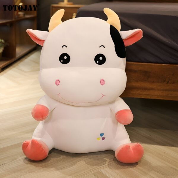 Une peluche blanche en forme de vache avec des détails roses et noirs. La vache a de grands yeux noirs, un sourire et une petite tache noire sur l'oreille. Elle est assise sur un plancher en bois.