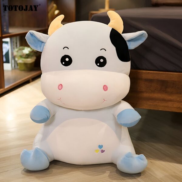 Peluche vache blanche avec des cornes jaunes, des yeux noirs et un sourire. La vache porte un petit cœur coloré brodé sur le ventre.