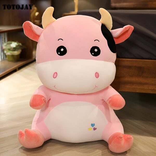 Une peluche rose en forme de vache avec de grands yeux noirs, des oreilles roses et une tache noire sur le côté. La peluche est assise sur un sol en bois et a un sourire amical.