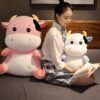Une jeune femme assise sur un lit avec deux peluches de vache, une rose et une blanche. La jeune femme porte une jupe à carreaux et un chemisier blanc. Elle lit un livre.