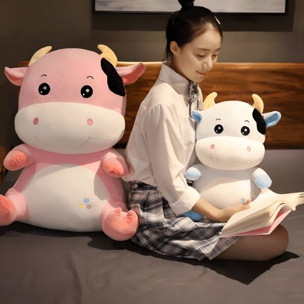 Une jeune femme assise sur un lit avec deux peluches de vache, une rose et une blanche. La jeune femme porte une jupe à carreaux et un chemisier blanc. Elle lit un livre.