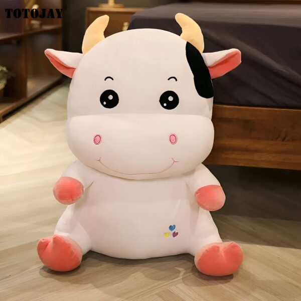 Une peluche de vache blanche avec des détails noirs, des yeux noirs et un sourire brodé. Elle a de petites oreilles roses et des pieds roses.