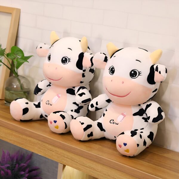 peluche-vache-jouet-enfant-bebe-cadeau-noel-2a Deux adorables peluches de vache noires et blanches assises sur une étagère en bois. Elles ont des visages souriants et portent le mot "Cow" sur leurs poitrines.